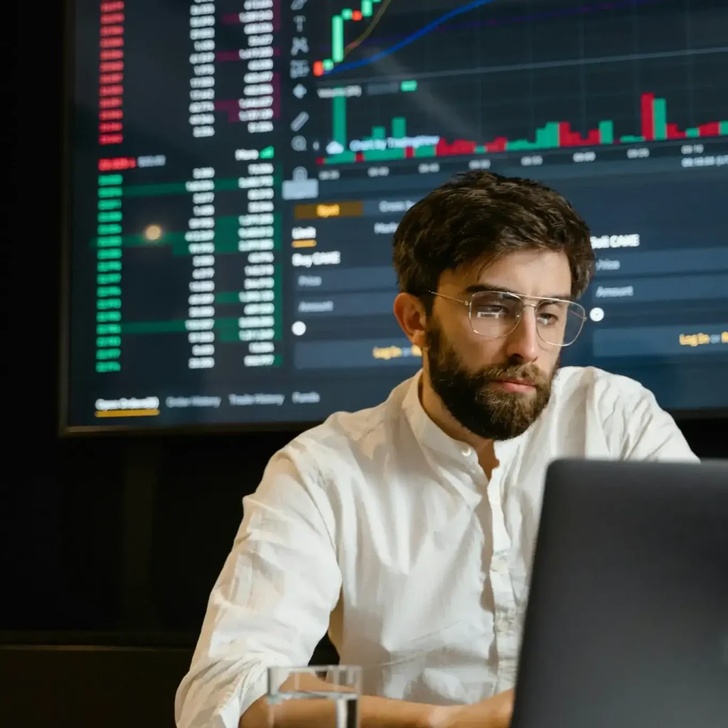 Professional man working on a laptop with financial charts displayed on a large screen behind him.